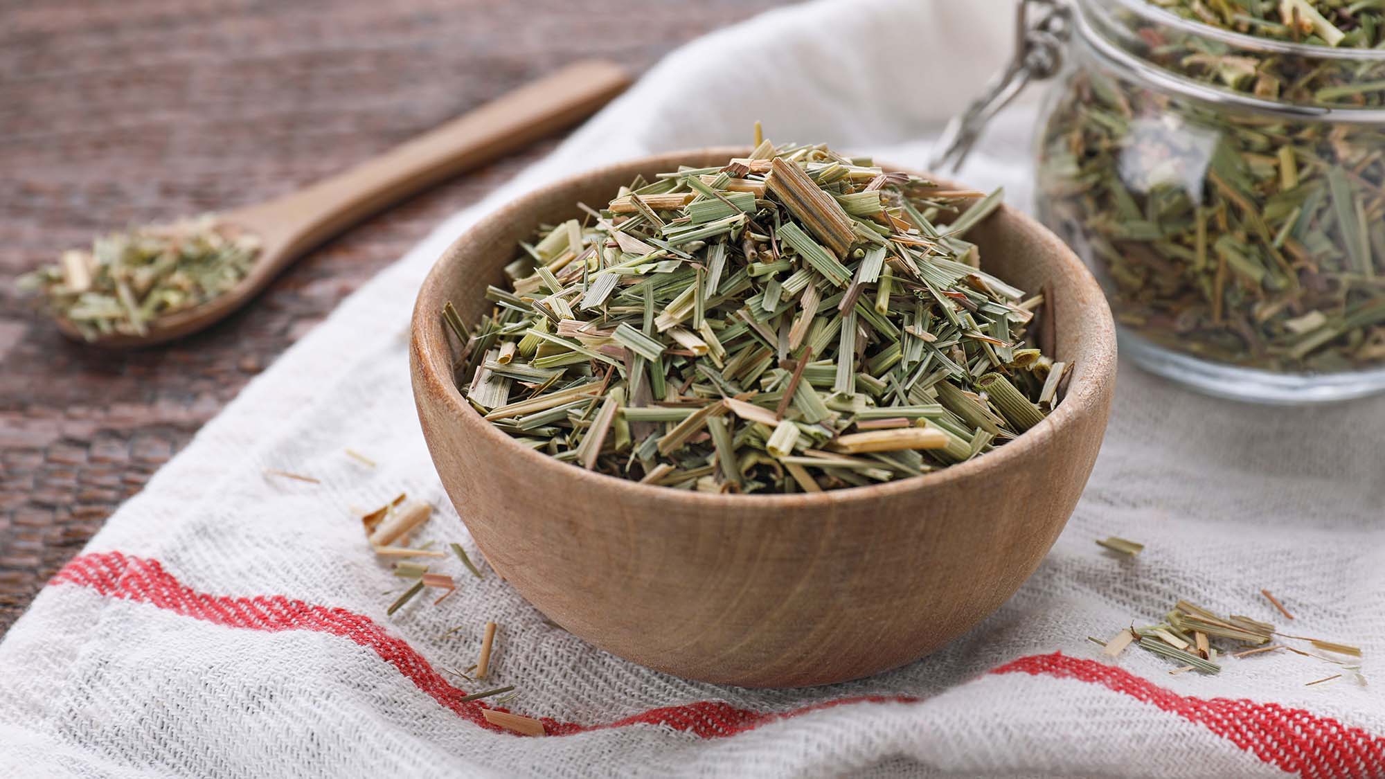 Wooden bowl filled with dried herbs on a cloth with a wooden spoon and jar in the background.